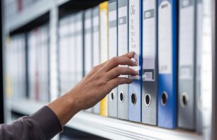 A person’s hand reaches precisely for a labeled blue binder among neatly aligned folders on an archive shelf, representing targeted document retrieval and systematic office organization.